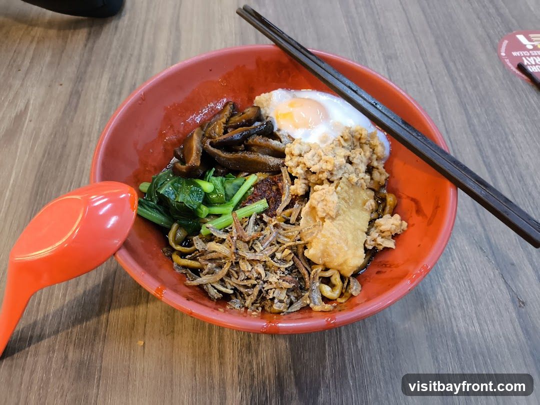A close-up shot of a bowl of noodles served with various toppings. The bowl is red and contains noodles, a fried egg, minced meat, shiitake mushrooms, leafy green vegetables, and crispy fried anchovies. A large orange spoon and a pair of dark chopsticks rest on the bowl. The table surface is made of light brown wood. A circular red and white sign is partially visible in the background.