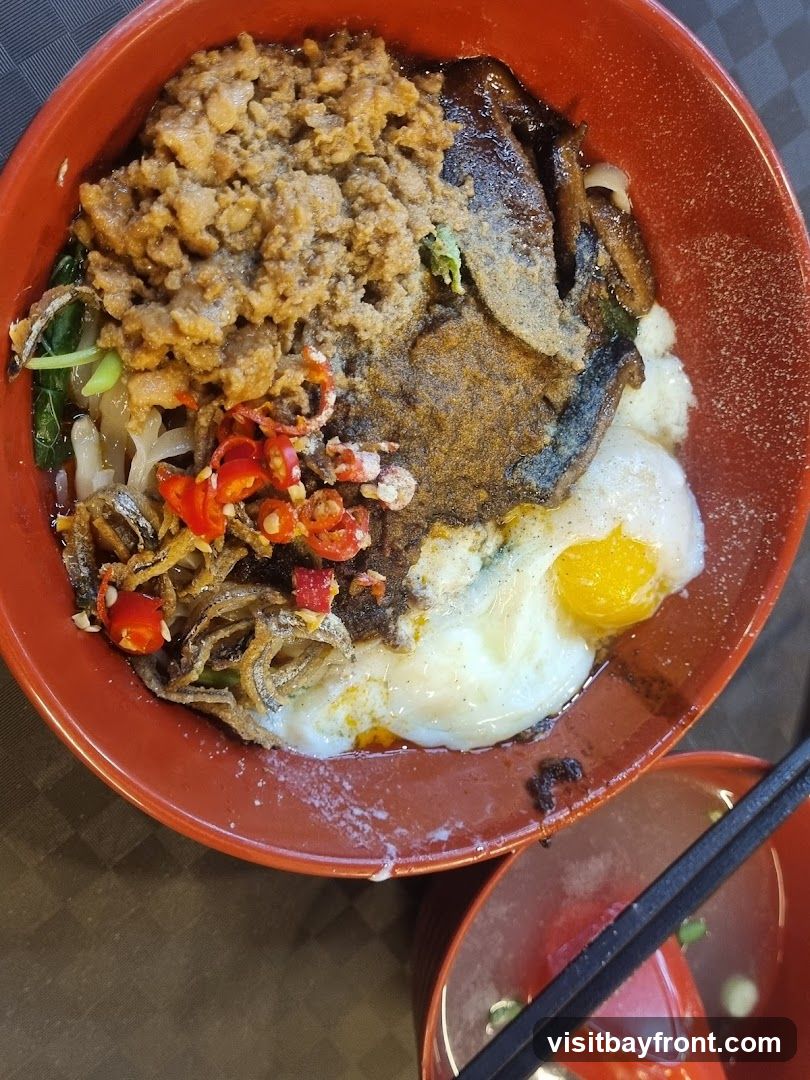 A close-up overhead view of a red bowl filled with noodles and various toppings. The toppings include minced meat, fried anchovies, sliced red chilies, a fried egg with a runny yolk, and what appears to be braised meat or mushrooms. A small bowl of soup and chopsticks are visible in the lower right corner.