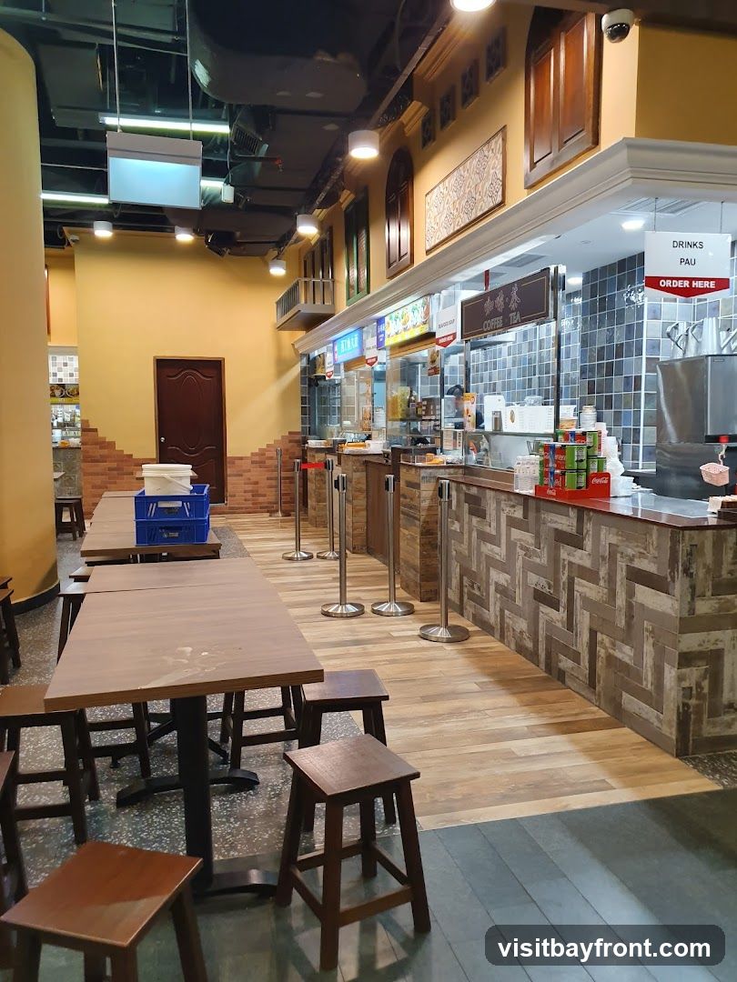 A food court with empty tables and chairs, and a counter area with food stalls. The counter has a tiled backsplash and a wooden front. There are signs above the counter indicating "COFFEE TEA" and "DRINKS PAU ORDER HERE". Canned drinks are visible on the counter. The seating area has wooden tables and stools.