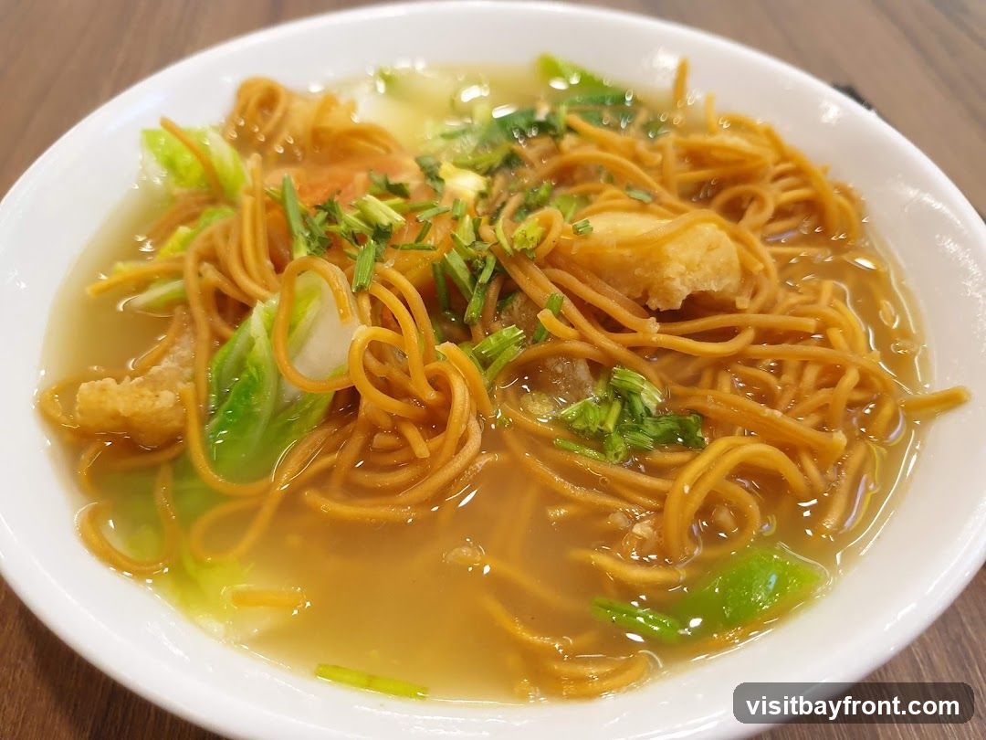 A close-up shot of a bowl of noodle soup. The soup contains thin, golden-brown noodles, pieces of fried tofu or fish, and chopped green vegetables, possibly cabbage and scallions. The broth is a light brown color.