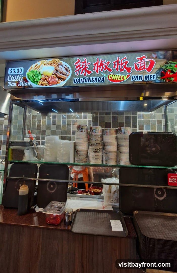 A sign above a food stall displays a picture of a bowl of Chilly Ban Mian, with the price $6.00. The sign also features Chinese characters and the text "DAMANSARA CHILLY PAN MEE". Below the sign, stacks of disposable cups and bowls are visible, along with serving trays and condiments. A person in an orange apron can be seen in the background.