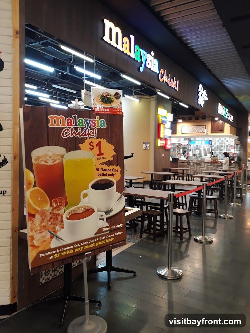 A food stall with the name "Malaysia Chiak!" is visible. A promotional sign displays drinks like Ice Lemon Tea, Lime Juice, and Hot Tea or Coffee for $1 with any meal purchase. The stall has seating areas with tables and stools.