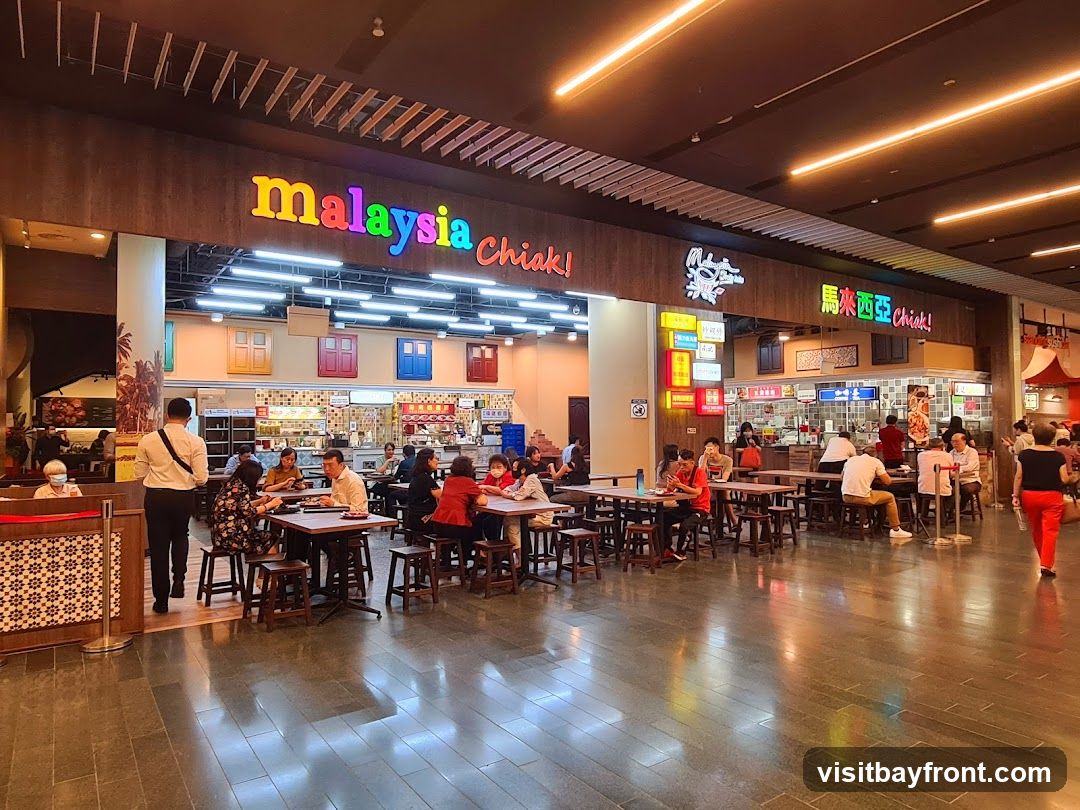 A bustling food court with multiple stalls and seating areas. The main stall features colorful signage that reads "Malaysia Chiak!". People are seen dining at tables and ordering food from various vendors. The overall atmosphere is lively and casual.
