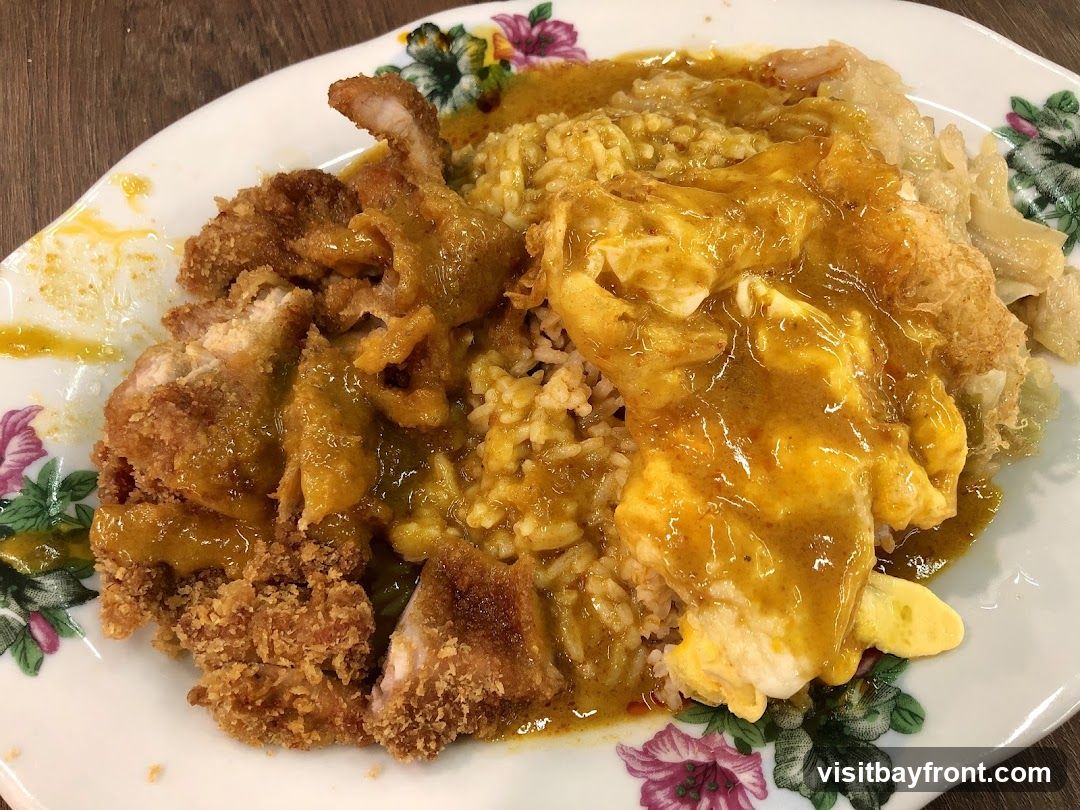 A close-up shot of a plate of curry rice. The dish features rice topped with crispy fried chicken pieces and a fried egg, all generously coated in a rich, yellow curry sauce. The plate has a floral pattern around the rim.