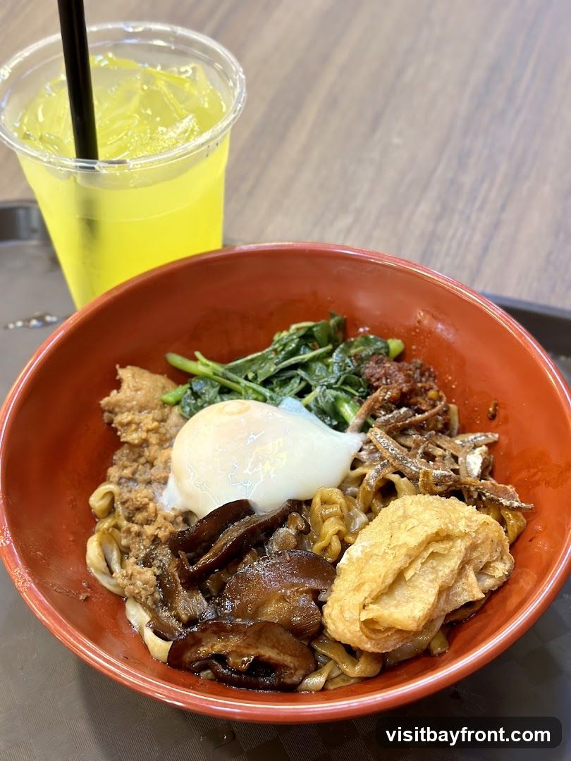 A close-up shot of a bowl of noodles topped with a poached egg, minced meat, shiitake mushrooms, greens, and fried anchovies. A glass of yellow beverage with ice and a straw is visible in the background.