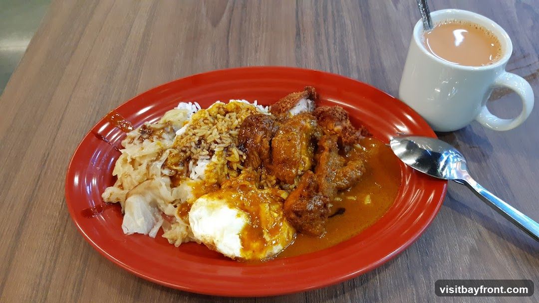 A red plate holds a serving of white rice topped with curry sauce, fried chicken pieces, and a fried egg. To the side, a white mug contains a beverage, likely tea, with a spoon. The meal is presented on a wooden table.