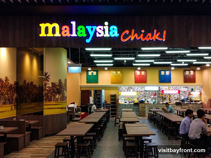 A brightly lit food court with a colorful sign that reads "malaysia chiak!". The interior features wooden tables and stools, with decorative colorful doors on the wall above the food stalls. A mural of a palm tree landscape adorns one of the walls. Several customers are seated at tables, and staff are visible behind the food counters.