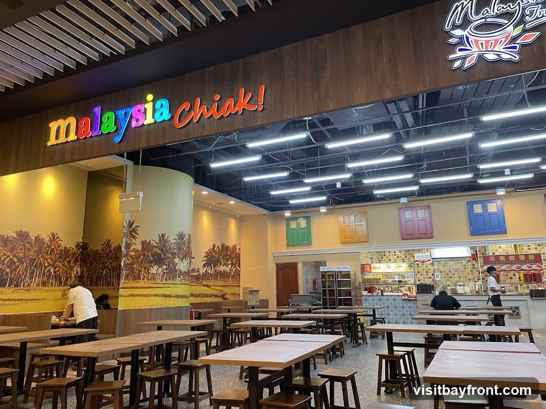 The interior of a restaurant with colorful signage that reads "Malaysia Chiak!". The walls are decorated with a mural of palm trees and rice paddies. There are many wooden tables and stools arranged throughout the dining area. In the background, there is a food counter with staff serving customers. Decorative windows in green, yellow, red, and blue are mounted on the wall above the counter.