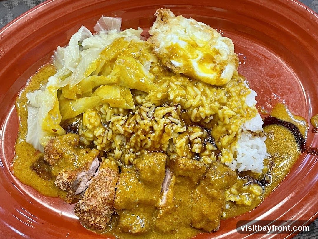 A close-up shot of a plate of curry rice. The dish includes white rice topped with a generous amount of yellow curry sauce, a fried egg with a runny yolk, braised cabbage, and pieces of breaded and fried meat, also covered in curry sauce. The food is served in a red, round bowl.