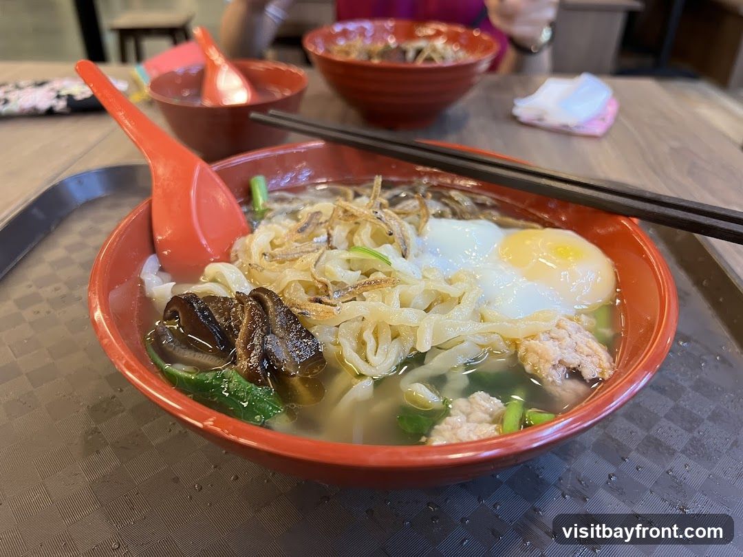 A close-up shot of a bowl of noodle soup. The soup contains flat noodles, a soft-boiled egg, minced meat, mushrooms, and greens. Fried anchovies are sprinkled on top. A red spoon and chopsticks rest on the edge of the bowl. In the background, another bowl of noodles and a person's hand are visible.