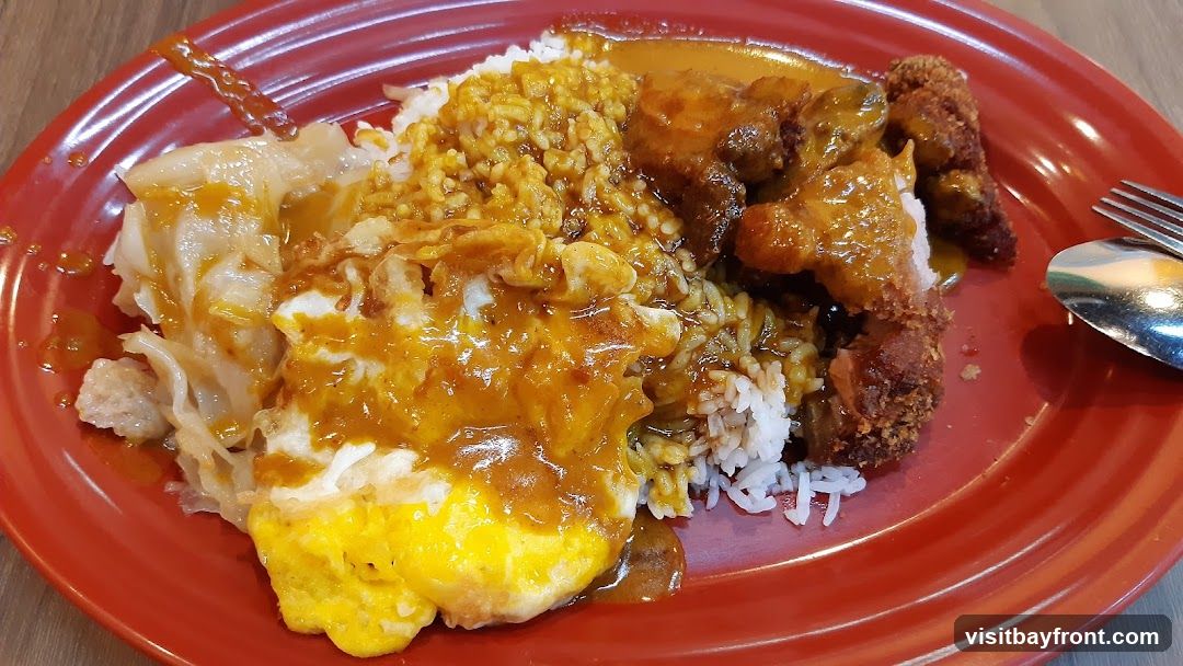 A close-up shot of a plate of curry rice. The dish includes white rice topped with a generous amount of curry sauce. On the side, there is a fried egg with a runny yolk, braised cabbage, and a piece of crispy fried chicken also coated in curry sauce. A spoon and fork are visible on the right side of the plate.