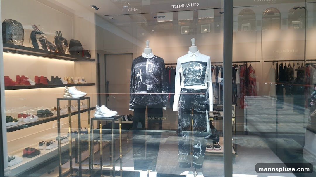 A fashion store window display featuring mannequins dressed in black and white graphic print outfits. The display also showcases various shoes and handbags on shelves and stands. The store's interior with racks of clothing is visible in the background. Reflections on the glass obscure some details.