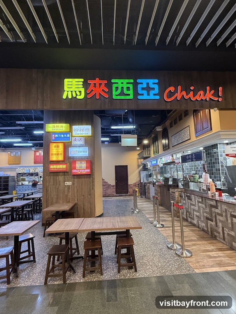 A food court with tables and chairs set up. A prominent sign with colorful Chinese characters and the English word "Chiak!" is visible above the entrance. Several smaller signs with food item names in Chinese and English are displayed on a pillar. The food stalls are visible in the background.