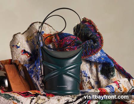 A dark green leather bucket bag with a criss-cross strap design and a single shoulder strap is displayed against a backdrop of textured fabric in vibrant blues, reds, and whites, with gold accents. The bag has the Alexander McQueen logo embossed on the lower front.