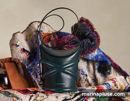 A dark green leather bucket bag with a criss-cross strap design and a single shoulder strap is displayed against a backdrop of textured fabric in vibrant blues, reds, and whites, with gold accents. The bag has the Alexander McQueen logo embossed on the lower front.
