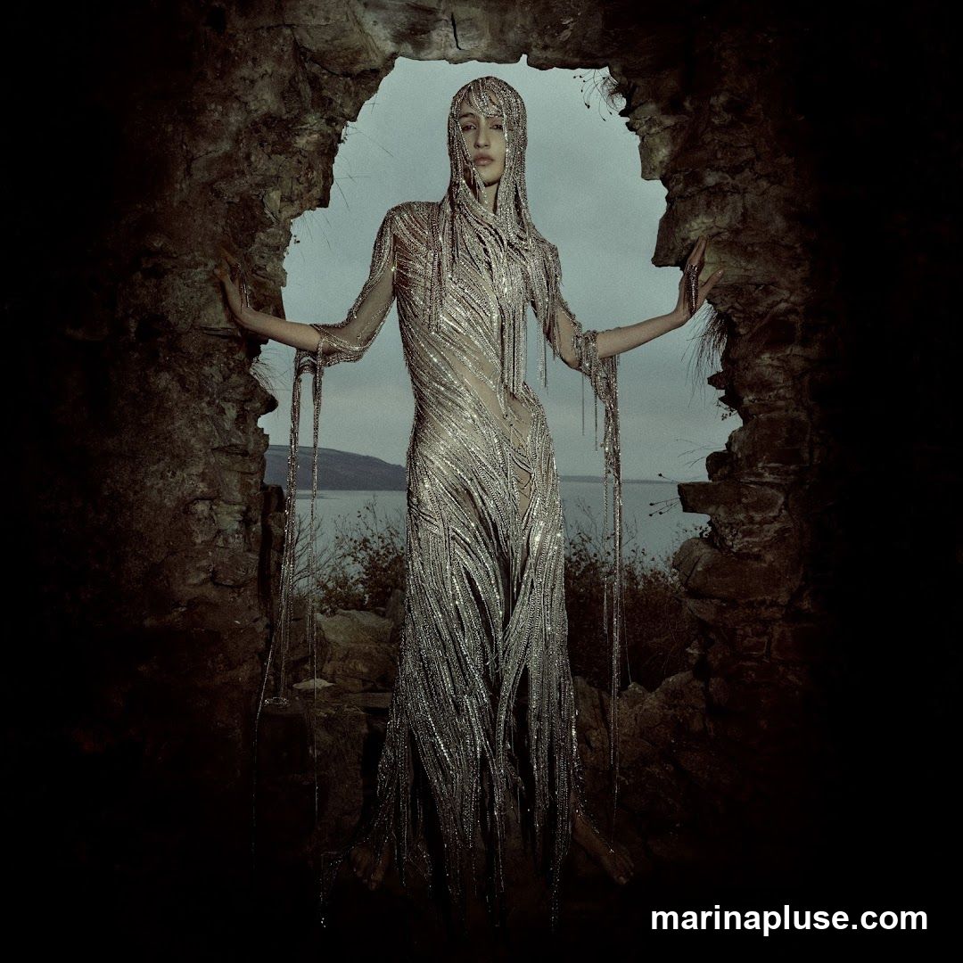 A woman stands within a stone archway, wearing an elaborate, shimmering gown made of metallic strands and beads. The gown has a hood that covers her head and cascades down her body, with long, dangling strands that reach past her knees. The background shows a body of water and a distant hill under an overcast sky. The overall mood is dramatic and ethereal.