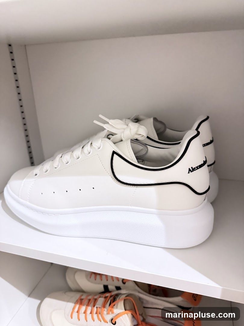 A pair of white Alexander McQueen sneakers with black trim and branding are displayed on a white shelf. Below them, another pair of sneakers with orange laces is visible.