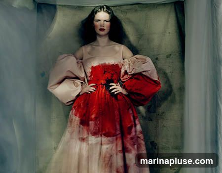 A woman with dark hair and bold red lipstick stands with her hands on her hips. She is wearing a voluminous gown with a light pink bodice and sleeves, and a skirt that transitions from light pink to white, heavily splattered with red paint or dye. The background is a textured, muted green fabric with some white drapery on the right.
