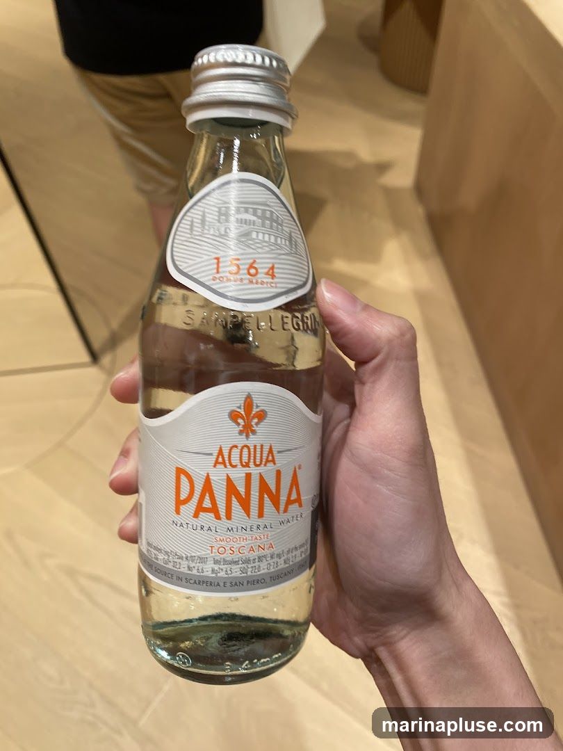A hand holds a glass bottle of Acqua Panna Natural Mineral Water. The label is white and orange and features the brand name "ACQUA PANNA" prominently. Below that, it reads "SMOOTH TASTE TOSCANA". The bottle has a silver cap. The background appears to be an indoor setting with light-colored wooden flooring.