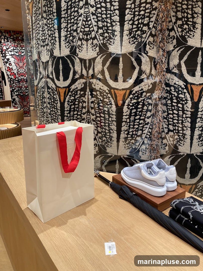 A white shopping bag with red handles sits on a wooden counter. Next to it, a pair of light grey sneakers with white soles and laces are displayed on a wooden stand. A black umbrella lies on the counter, and a folded black and white patterned fabric is visible to the right. The background features a striking black and white patterned wallpaper with orange accents, possibly depicting feathers or stylized insects.