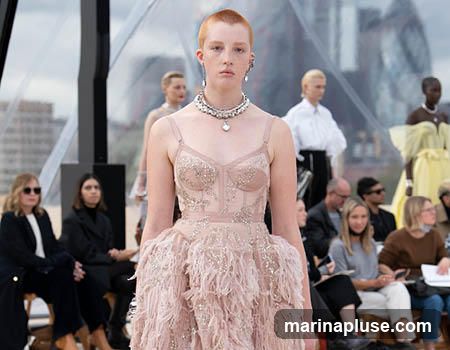 A fashion show with models on a runway and an audience seated. The main model in the foreground wears a light pink, embellished corset dress with feathered detailing. Other models in the background are also wearing elaborate outfits. The audience members are dressed in casual to semi-formal attire. The setting appears to be outdoors with a blurred background of buildings and a translucent roof structure.