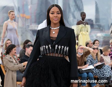 Naomi Campbell walks down a runway during a fashion show. She is wearing a black blazer with intricate silver embellishments and a black feathered skirt. Other models and audience members are visible in the background.