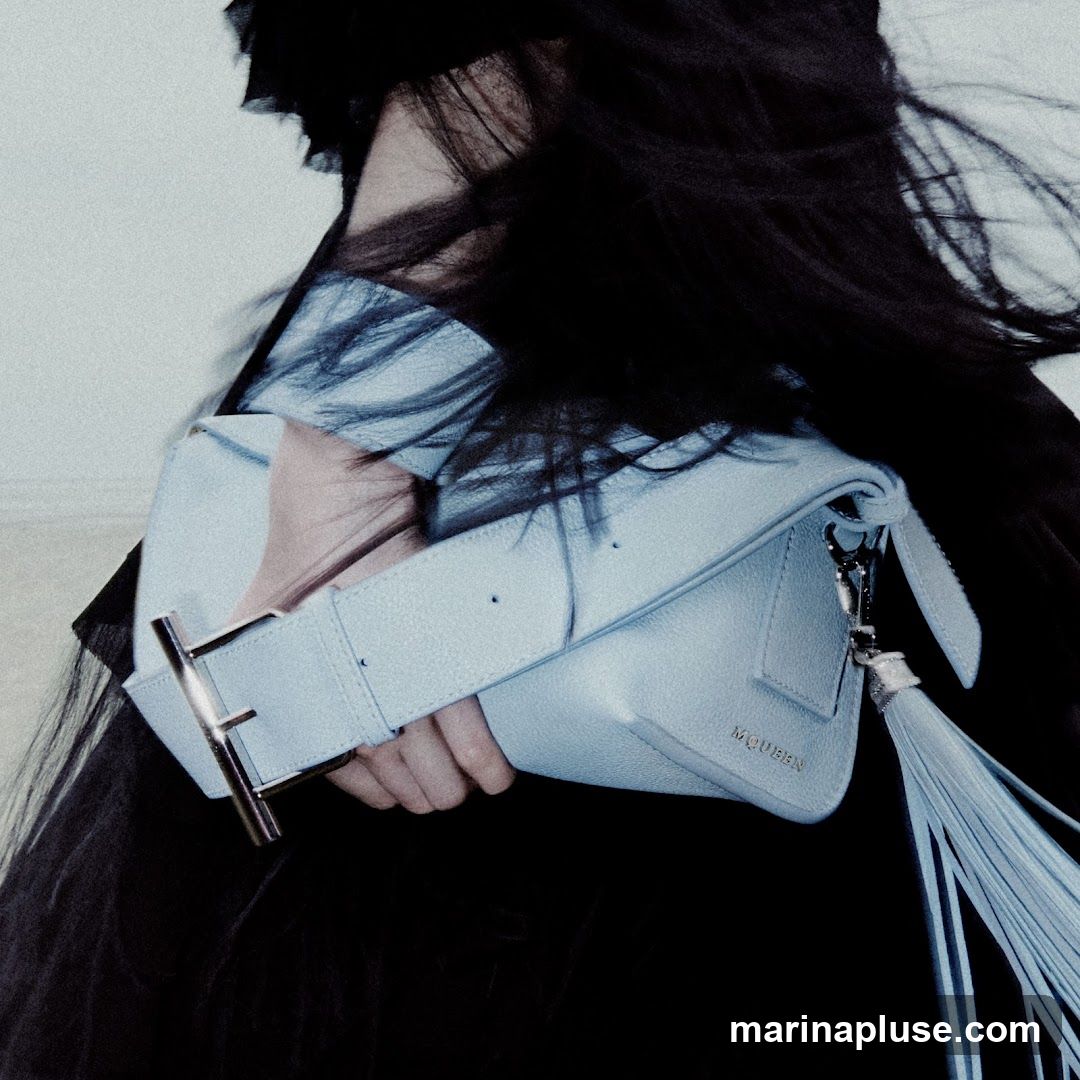 A person with long, dark hair holds a light blue leather handbag. The bag has a wide strap with a silver buckle and a long tassel. The brand name "MCQUEEN" is embossed on the front of the bag. The person is wearing a black garment.