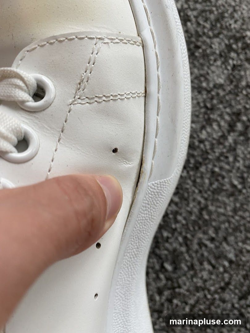 A close-up view of a white leather sneaker, showing stitching details, eyelets for laces, and small perforations. A thumb is visible, pointing to a slight separation in the sole. The background is a textured grey carpet.