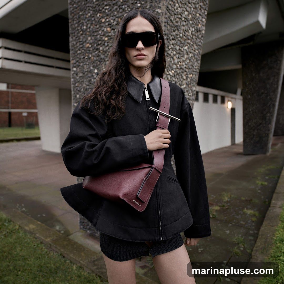 A woman wearing sunglasses, a black jacket, and shorts, posing with a burgundy handbag. The background features brutalist architecture with concrete pillars and textured walls.
