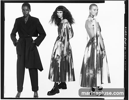 Three models are posing in a studio setting. The model on the left wears a black cropped blazer with wide-leg trousers. The middle model wears a flowing dress with an abstract print. The model on the right also wears a dress with a similar abstract print, featuring an open back. The image is a black and white photograph with a film border.