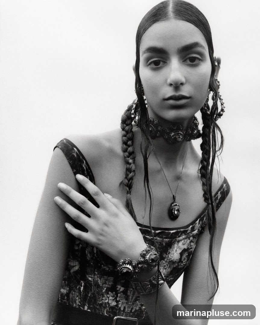 A black and white portrait of a young woman with dark hair styled in two braids. She is wearing a patterned dress, a choker necklace, a pendant necklace, large hoop earrings, and a bracelet. Her left hand rests on her right arm. The background is plain white.
