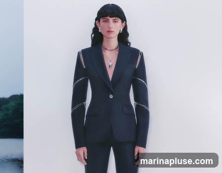 A woman with dark hair and bangs stands against a white background, wearing a tailored black blazer with zippers on the sleeves and a matching pair of pants. She is also wearing a necklace with a pendant and earrings. To the left of the woman is a blurred image of a body of water and trees.