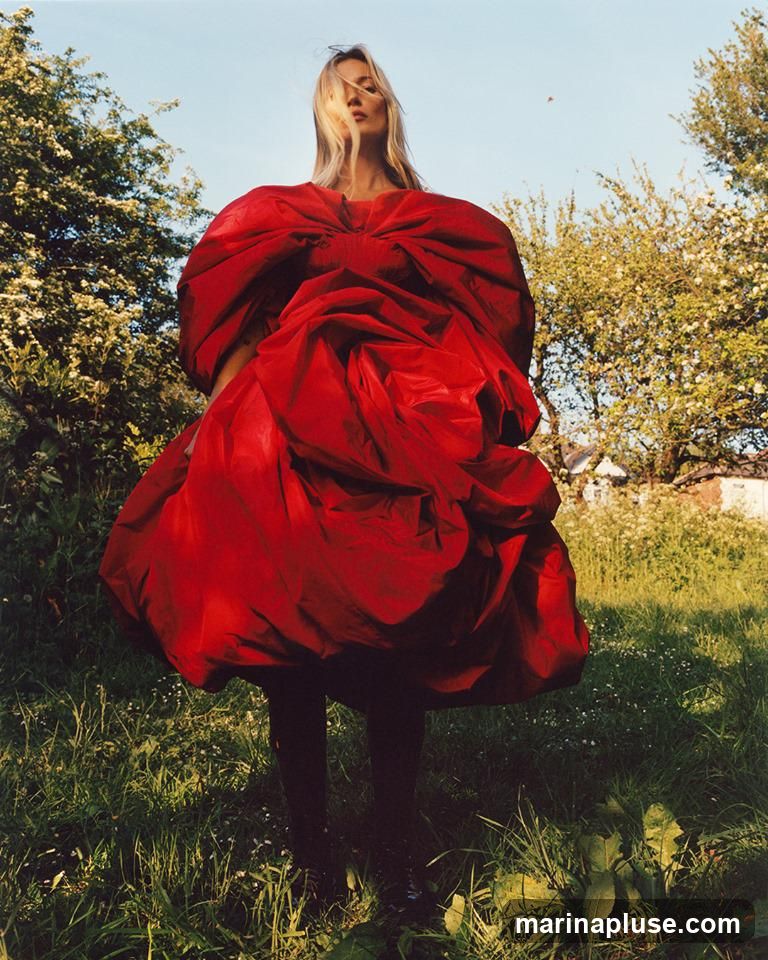 A woman with blonde hair stands in a grassy field, wearing a voluminous red dress with a large bow detail at the neckline. She is facing forward, with her face partially obscured by her hair. The background features trees with white blossoms and a clear blue sky.