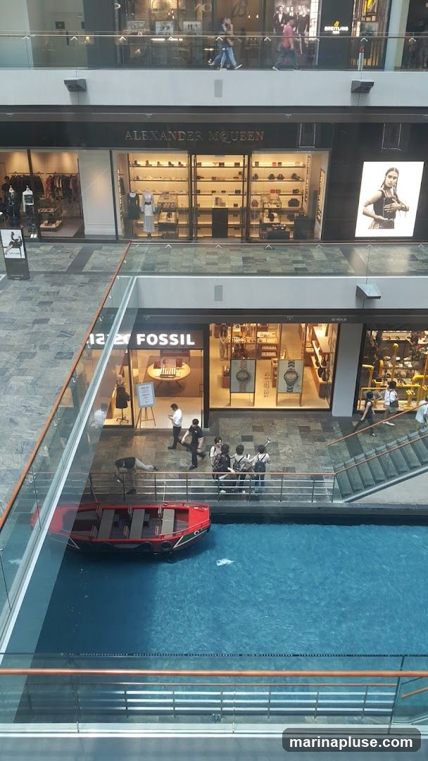 An overhead view of a multi-level indoor shopping mall. A red boat is floating in a blue canal in the foreground. Shops are visible on the upper levels, including Alexander McQueen and Fossil. People are walking on the upper levels and standing near the canal.