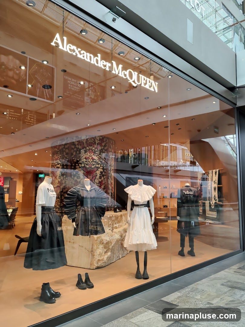 The storefront of an Alexander McQueen boutique, featuring a large glass window displaying mannequins dressed in the brand's fashion. The store's name is prominently displayed in illuminated white lettering above the window. Inside, the store appears to be luxuriously appointed with displays of handbags and other merchandise.