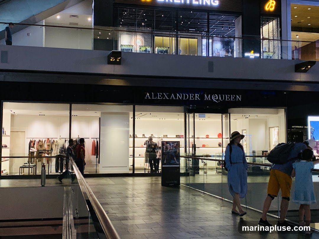 The exterior of an Alexander McQueen store is visible, with large glass windows displaying clothing and accessories. The store's name is prominently displayed in large letters above the entrance. People are seen walking past the store and inside. In the background, another store with the name "BREITLING" is visible.