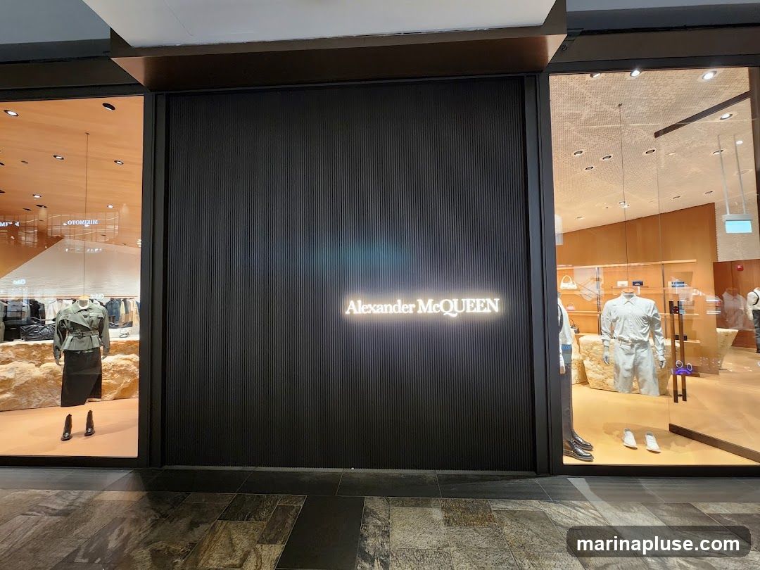 The exterior of an Alexander McQueen store. The storefront features a dark, ribbed facade with the brand name illuminated in white neon. To the left and right are large glass windows displaying mannequins dressed in the brand's clothing and accessories. The interior of the store is visible through the windows, showcasing shelves stocked with merchandise.
