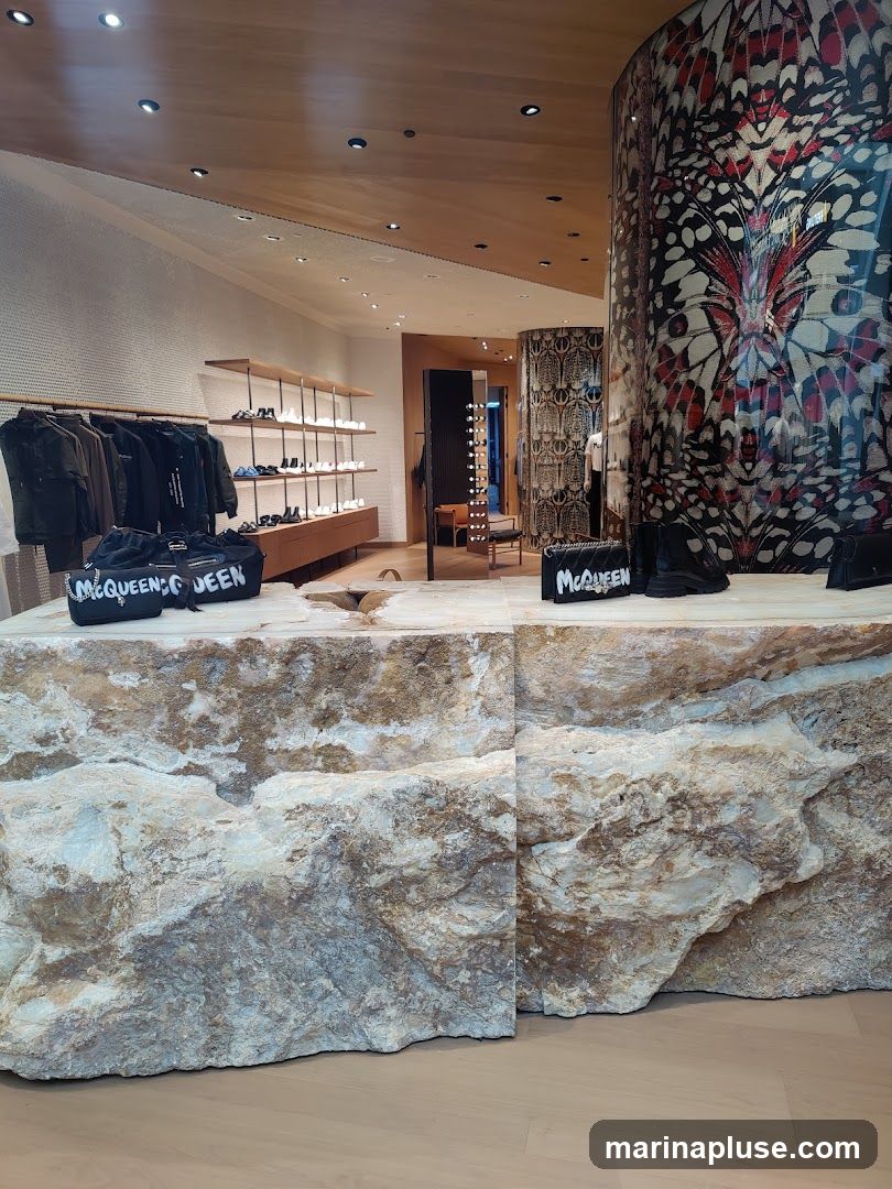 The image shows the interior of a high-end retail store. In the foreground, a large, rough-hewn stone counter displays several black handbags with white lettering that reads "MCQUEEN" and a pair of black boots. Behind the counter, racks of clothing are neatly displayed, and shelves showcase various shoes. A striking, large-scale butterfly-patterned pillar dominates the right side of the frame, adding a dramatic visual element to the space. The overall aesthetic is modern and luxurious, with clean lines and natural materials.