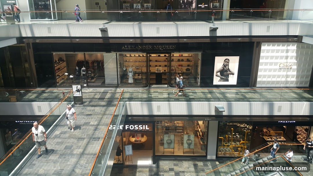 An interior view of a multi-level shopping mall, showcasing various high-end retail stores. Prominently featured are the storefronts of Alexander McQueen and Chloe. Other visible stores include Fossil and a store with the name 'FALZION'. People are seen walking on the walkways and escalators, browsing the shops, and interacting within the mall environment. The architecture is modern, with glass railings and a spacious, well-lit atrium.