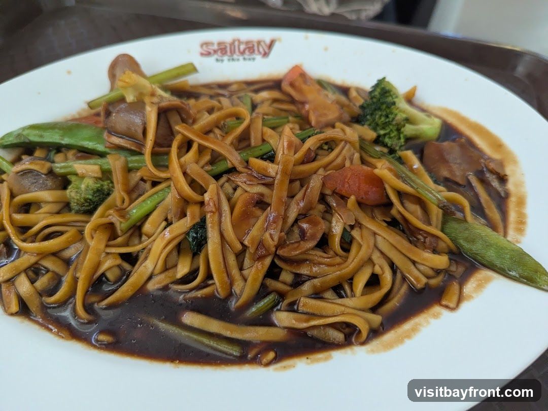 A plate of noodles with mixed vegetables, including broccoli, snap peas, and mushrooms, in a dark sauce. The logo "satay by the bay" is visible on the rim of the plate.