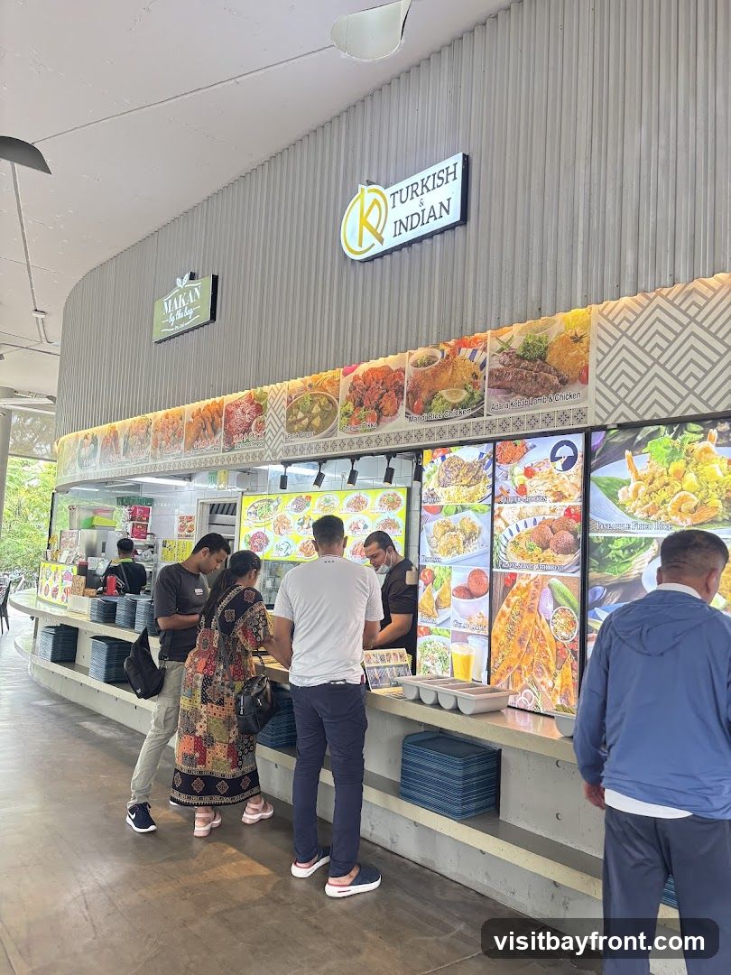 A food stall with a sign that reads "TURKISH & INDIAN" and a logo with the letter R. Below the sign are large colorful pictures of various dishes. People are standing in line to order food.