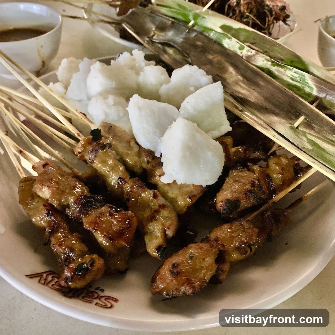 A close-up shot of a plate of chicken satay served with rice cakes. The satay skewers are arranged on a white plate with a logo that reads 'Satey by...'. The chicken pieces are grilled and glazed, with some char marks visible. Several white, cubed rice cakes are placed in the center of the plate. A small bowl of dipping sauce is visible in the upper left corner, and some grilled items wrapped in leaves are in the background.
