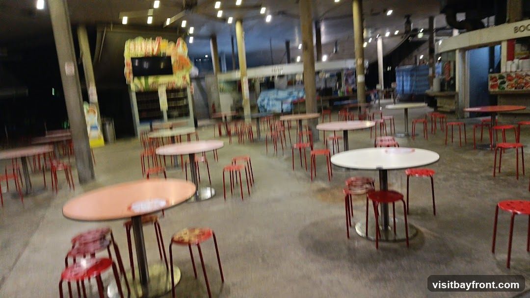 An outdoor seating area with numerous tables and red stools. The tables are round and vary in color from pink to white. The area appears to be part of a food court or market with various stalls visible in the background. Some stalls have signage, including one with colorful graphics and a screen. The lighting suggests it is evening or night.