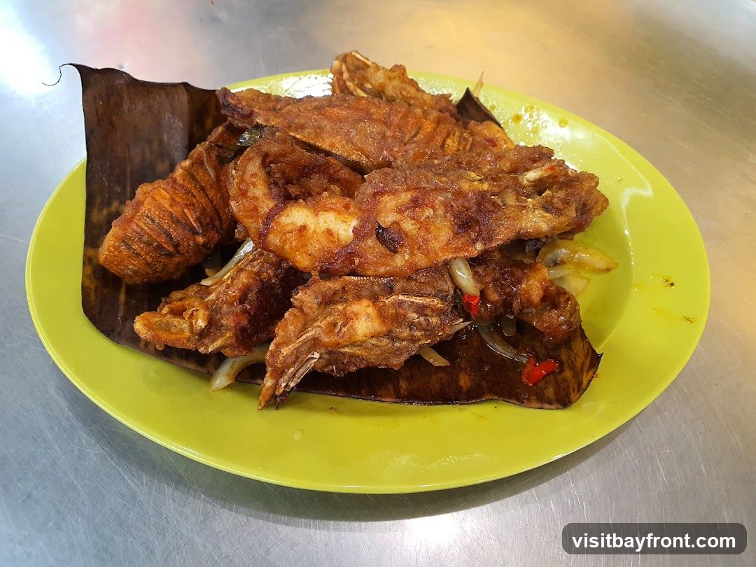 A bright yellow plate holds a serving of fried fish pieces, some of which are whole fish. The fish is golden brown and appears crispy. It is served on a bed of dark green banana leaves and garnished with sliced onions and red chili peppers. The plate is placed on a metallic surface.