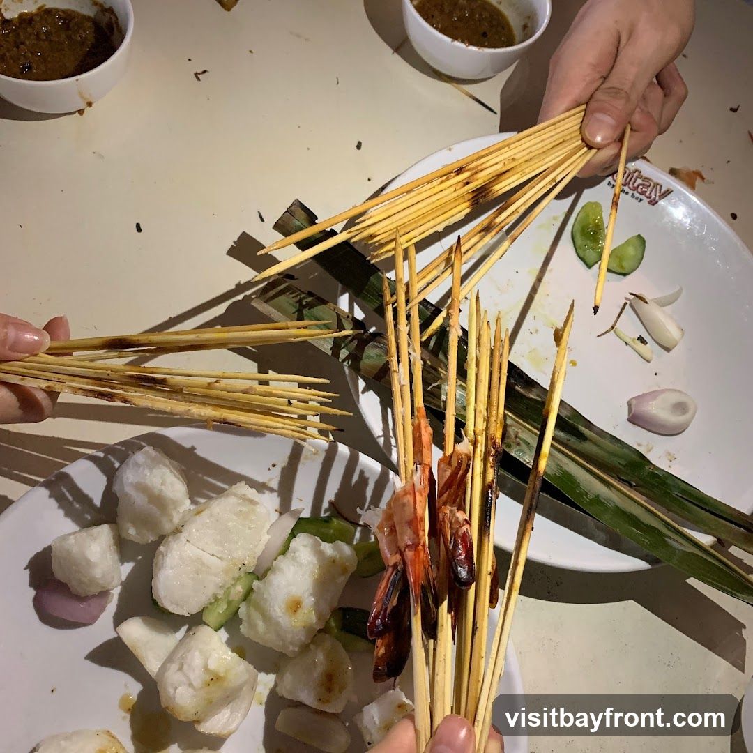 A close-up overhead shot of several plates of food, with hands holding skewers of grilled shrimp and other meats. The skewers are made of bamboo and have been charred from grilling. The plates contain rice cakes, cucumber slices, and red onion. Two small bowls of dipping sauce are visible in the background. The overall impression is of a shared meal, likely satay.