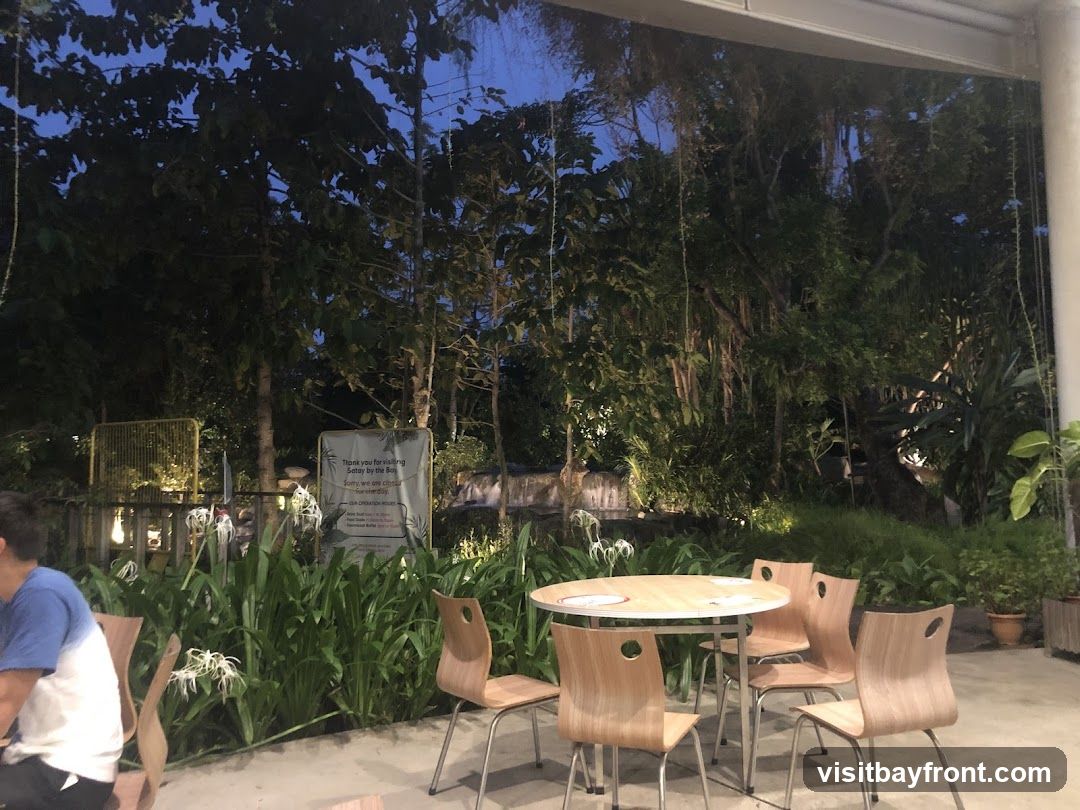 An outdoor seating area with wooden chairs and a round table is set against a backdrop of dense, lush greenery and trees. The lighting suggests it is dusk or evening. A sign is visible in the background with text that appears to be operational hours and a thank you message.