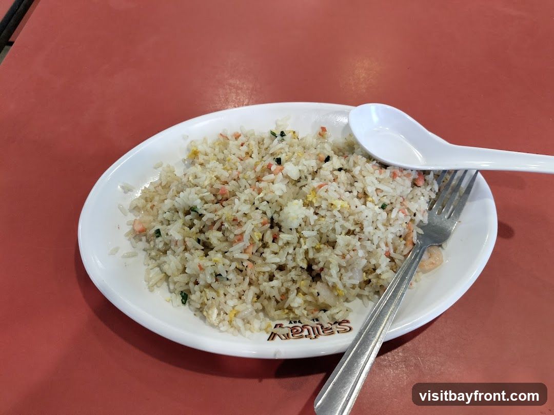 A plate of fried rice with visible pieces of shrimp and egg, served with a spoon and fork on a red table. The plate has a logo that reads "Saitay's" on the bottom.