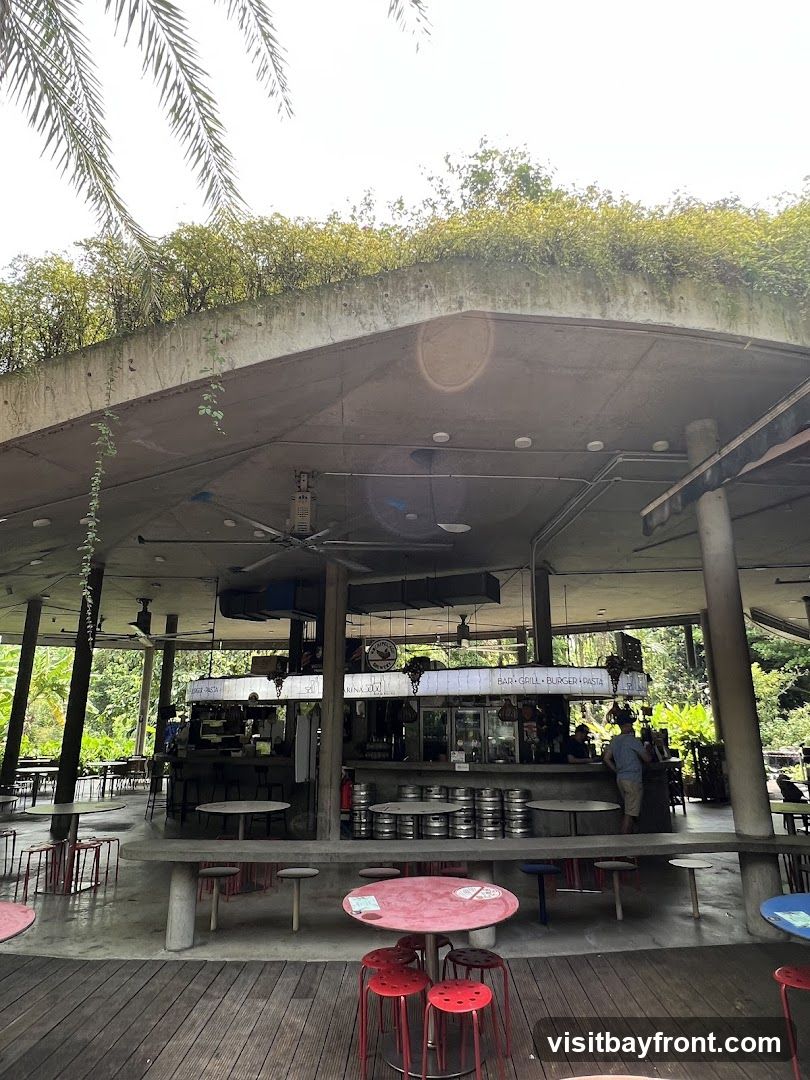 An open-air bar and grill area with a concrete roof covered in greenery. Tables and red stools are arranged on a wooden deck in the foreground. In the background, a bar area is visible with kegs, refrigerators, and staff serving customers. Signage above the bar indicates "BAR GRILL BURGER PASTA".