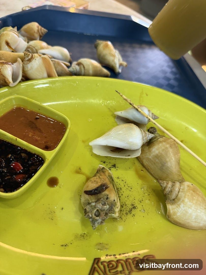 A bright yellow plate holds several cooked sea snails, some on skewers, and two small dishes of dipping sauce. One sauce is a reddish-brown, and the other is dark with visible chili pieces. In the background, a blue tray holds a pile of more sea snails. The plate has a partially visible logo that reads 'Satay'.