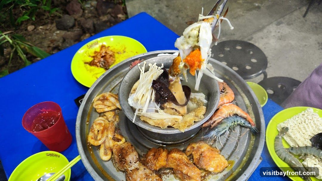 A close-up view of a hot pot meal with various ingredients. In the center, a crab is being lifted, revealing its roe and innards, surrounded by enoki mushrooms and other seafood. Around the pot, there are grilled meats, shrimp, and a plate of instant noodles. A red cup with a beverage and a green bowl with chopsticks are also visible on a blue table.
